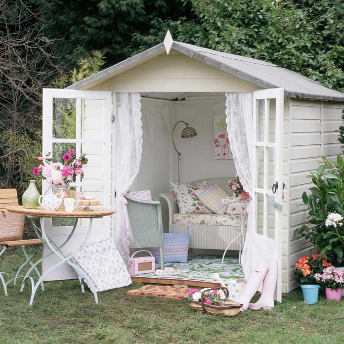 White summer house shed with doors open, lace curtains, floral cushions and Lloyd Loom furniture