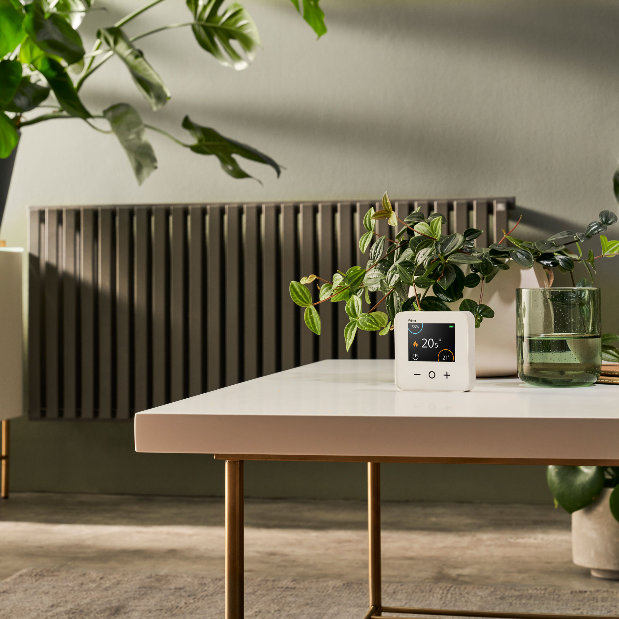 smart thermostat on a table in a living room with a radiator in the background and plenty of greenery from potted plants