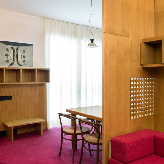 Dining area with wooden chairs surrounded by built-in wooden shelving and magenta carpet in living room of the Casa Lana installation at Triennale di Milano, Italy