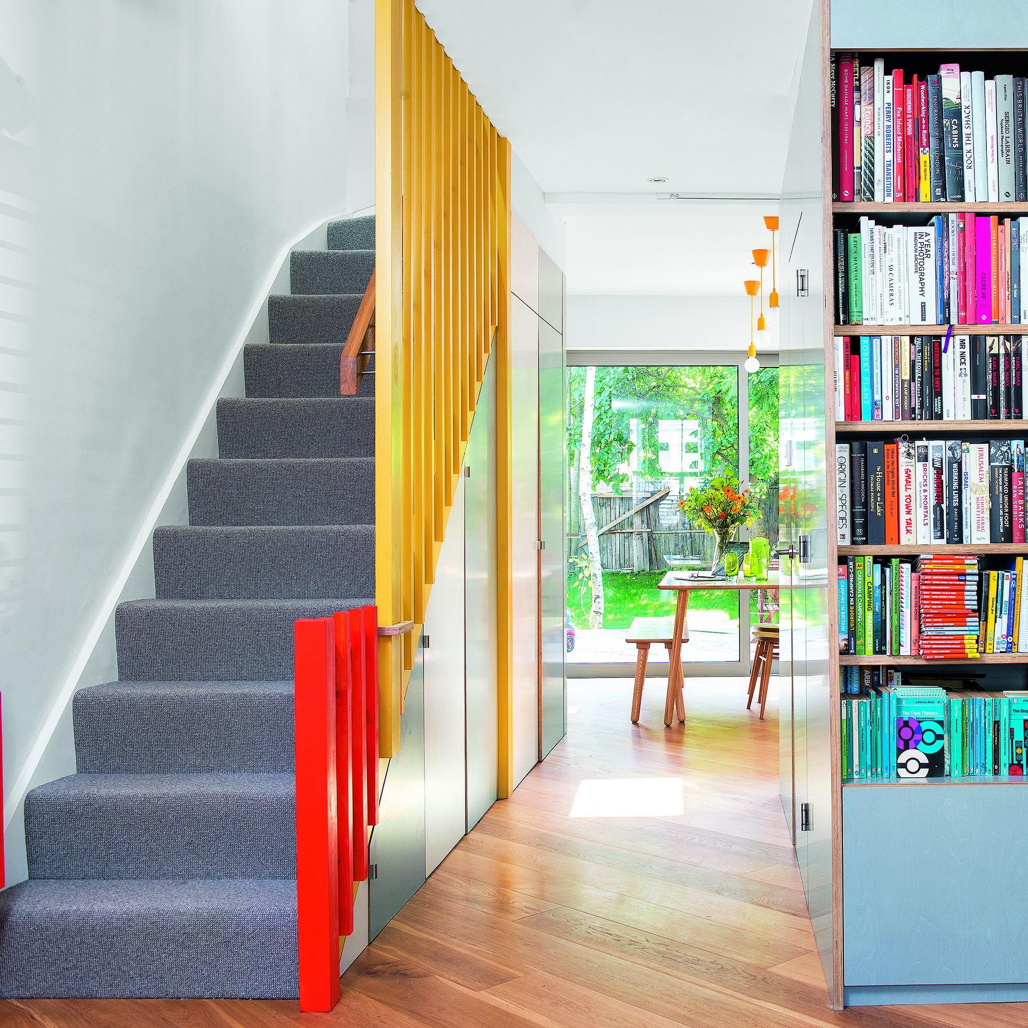 Hallway with storage and bookcase and doors painted in duck egg blue, with staircase opposite with grey stair carpet and yellow and orange painted bannisters