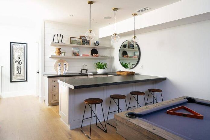 Behind an oak pool table, Iron and wood backless barstools sit at a light gray basement bar island lit by glass and brass mini pendants. A round mirror hangs adjacent to stacked light gray floating shelves mounted to a white shiplap wall over light gray shaker cabinets donning oil rubbed bronze hardware and a black countertop.