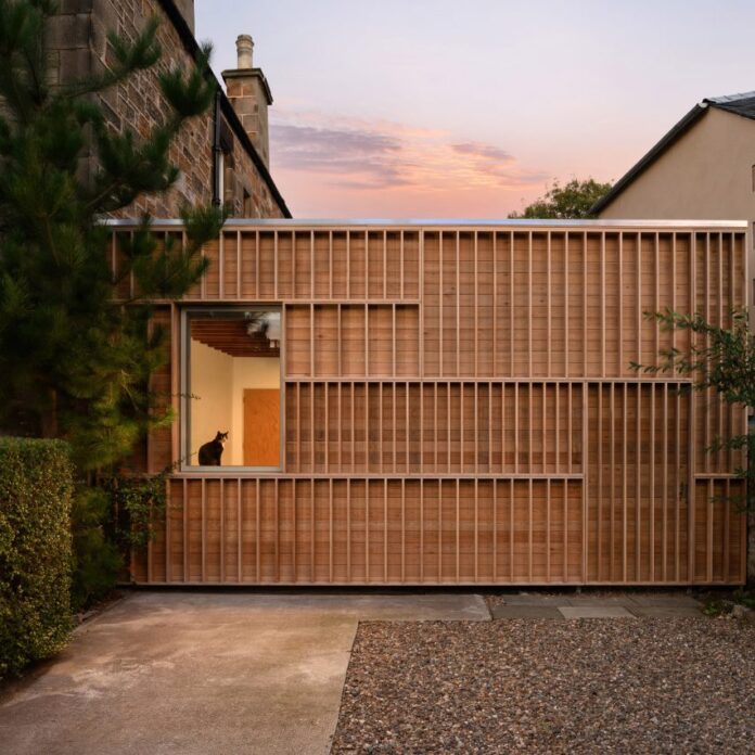 Photograph showing a timber-clad structure on the side of a house at dusk/dawn with a cat in the window