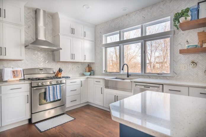 Stacked white shaker kitchen cabinets with a large window over the sink, marble herringbone tile, and warm wood floating shelves