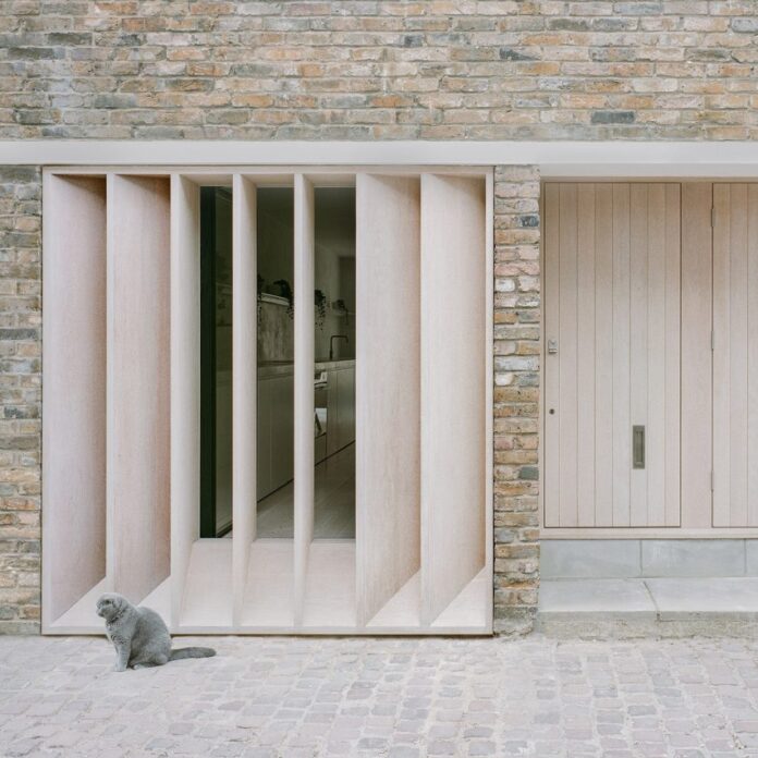 Exterior of a brick mews house with a timber door, timber louvres in front of a large window and a grey cat