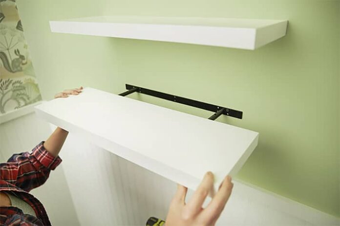 Man installing two white floating shelves on drywall.