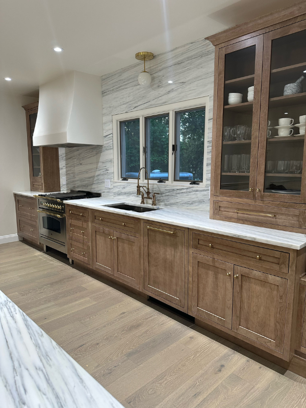 Medium-brown inset cabinets with furniture-style tall glass wall cabinets sitting on a white stone countertop with an artisan white kitchen hood