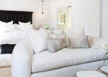 Under a dark stained wood ceiling beam, a white blush curved sofa sits on a Moroccan style wood rug in a bedroom sitting area featuring a white and gold accent table. Behind the sofa, on a white wool rug, a Restoration Hardware 17th C. Castelló Bed With Footboard is lit by a blow glass pendant hung over a nightstand.