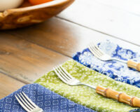 4 finished blue, green and white napkins folded and lined up on a table with a fork on each.