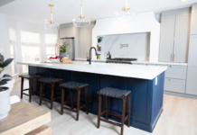 Large one-wall kitchen design with tall light gray shaker kitchen cabinets and gold cabinet hardware with a long dark blue kitchen island and white quartz countertops